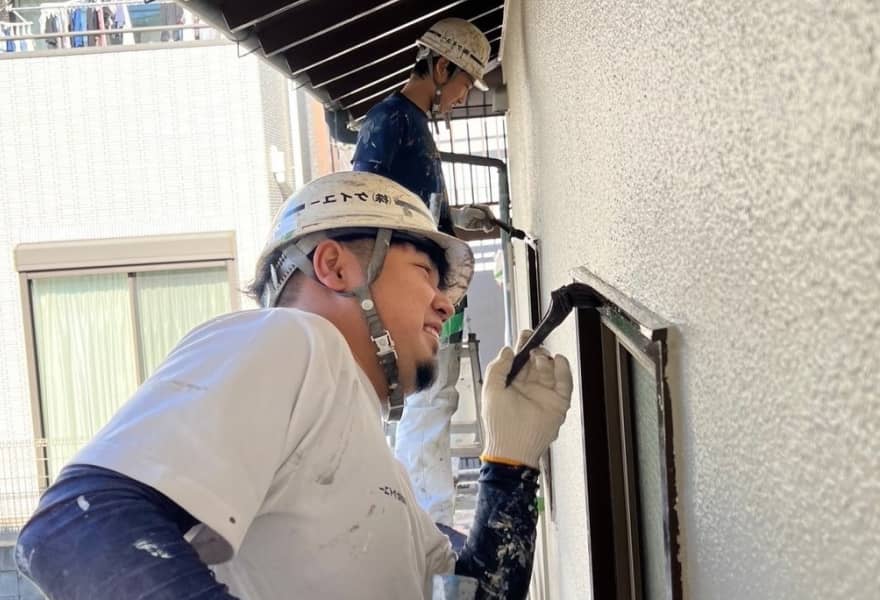 建物の外壁を修理・塗装している作業員が2人写っています。1人は窓枠を塗装し、もう1人は脚立に上がって作業をしています。作業現場は晴れた日で、背景には隣接する建物が見えます。