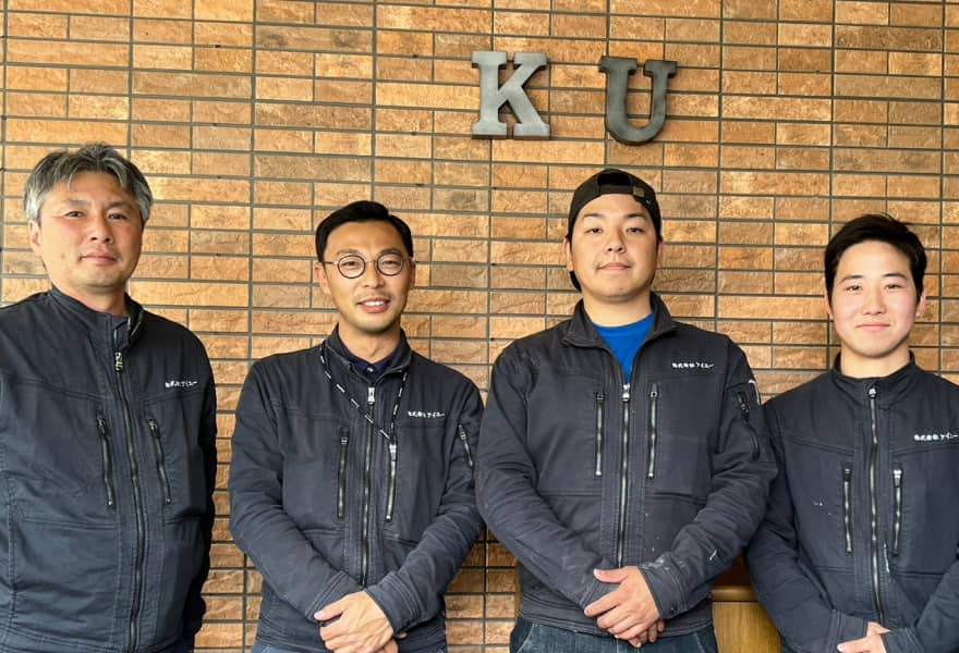 建物の前で撮影された、4人の男性スタッフ。背景にはレンガ調の壁と「KU」の文字が見える