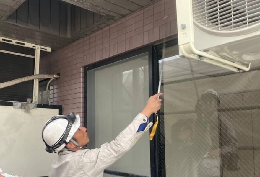 白いヘルメットを被った作業員が、エアコン室外機周辺で棒状の道具を使って調整作業をしている