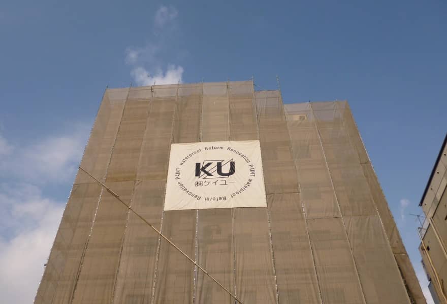 青空を背景に、工事用の足場に囲まれた建物。中央には「KU 株式会社ケイユー」のロゴ入り幕が取り付けられている